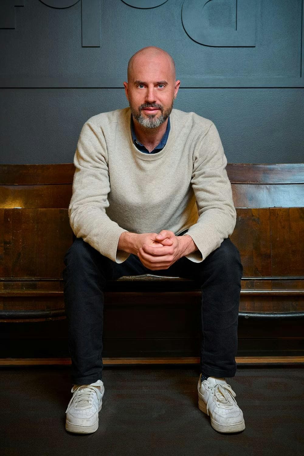 Full-body shot of Eric Olofsson, a white man with a beard wearing a light grey sweater, dark jeans, and white sneakers. He sits on a chair against a dark gray wall.