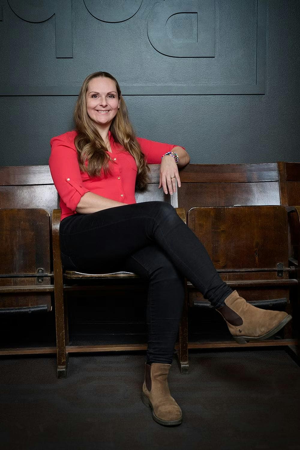 Full-body shot of Caroline Lengholm, a white woman with long auburn hair and wearing a red shirt and black pants. She is sitting confidently on a chair against a dark gray wall.